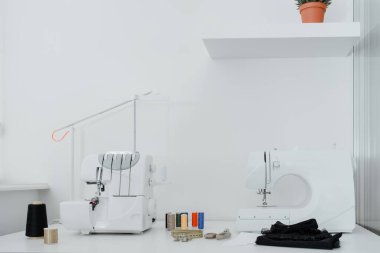 Creativ tailor workplace. Close up sewing and overlock machines, spool of thread, scissors, centimeter ribbon and pieces of black material on the table