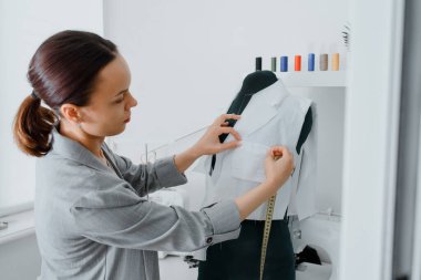 Young woman tailor is measuring with a centimeter ribbon a future shirt on a mannequin in her mini workshop