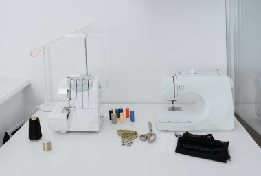 Creativ tailor workplace. Close up sewing and overlock machines, spool of thread, scissors, centimeter ribbon and pieces of black material on the table