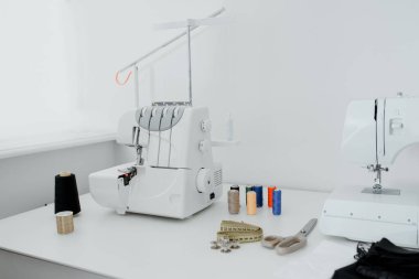 Creativ tailor workplace. Close up sewing and overlock machines, spool of thread, scissors, centimeter ribbon and pieces of black material on the table