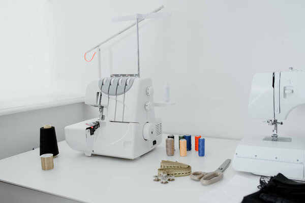 Creativ tailor workplace. Close up sewing and overlock machines, spool of thread, scissors, centimeter ribbon and pieces of black material on the table