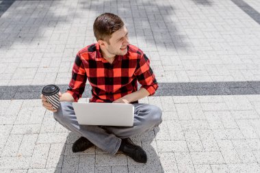 Genç serbest çalışan adam dışarıda bilgisayar başında çalışıyor ve kahve içiyor. O, sokak zemininde oturuyor. Her yerde çalışıyor.