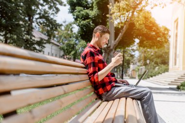 Mutlu genç serbest çalışan adam dışarıda bilgisayar başında çalışıyor ve kahve içiyor. O, sokak tahtası sandalyesinde oturuyor. Her yerde çalışıyor.