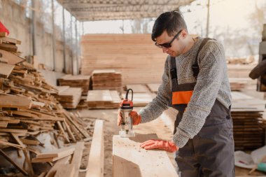 Ahşap atölye deposundaki genç marangoz ahşap kereste yığınından yapılmış ahşap bir kereste / tahtanın üzerinde değirmen makinesi kullanıyor..