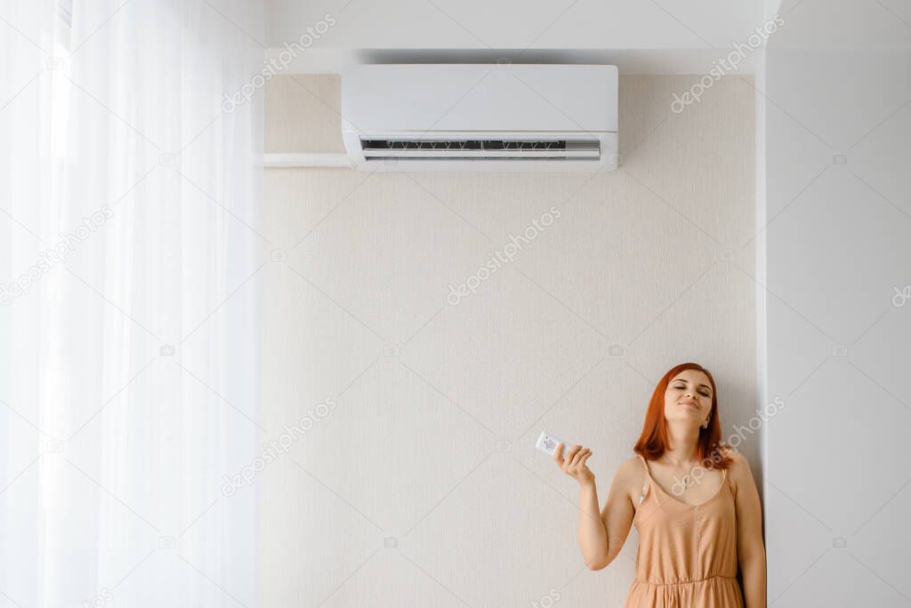 Young beautiful woman in her white apartment turns on the air conditioner and enjoys the cool. Fight with heat concept