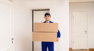 Mavi tulumlu gülümseyen genç kurye iki karton kutuda bir paket teslim ediyor. Kapılı beyaz arkaplan