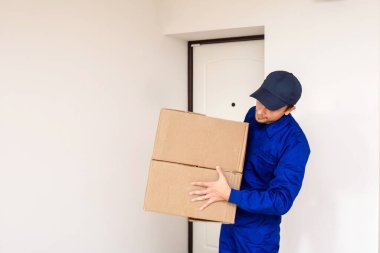 Mavi üniformalı genç teslimatçı müşteri için bir paket yolluyor. Kapılı beyaz arka plan. Metin için boşluk kopyala