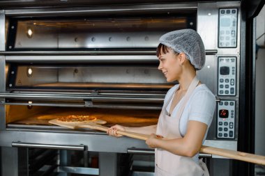 Kafkasyalı genç fırıncı elinde taze pizzayla ahşap kabuğu tutuyor ve bir fırında pişiriyor. Lezzetli pizza fabrikası konsepti.