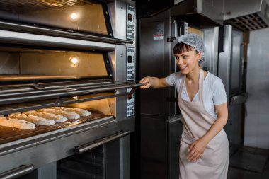 Genç gülümseyen beyaz kadın fırıncı, fırın fabrikasındaki elektrikli fırında çalışan ekmek fırınına bakıyor. Fransız ekmeği fırını konsepti.