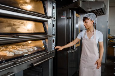 Genç beyaz kadın fırıncı ekmek fırınına bakıyor ve bir fırın fabrikasının elektrikli fırınını açıyor. Fransız ekmeği fırını konsepti. 