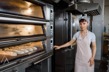 Genç beyaz kadın fırıncı ekmek fırınına bakıyor ve fırın fabrikasındaki elektrikli fırını açıyor. Fransız ekmeği fırını konsepti.