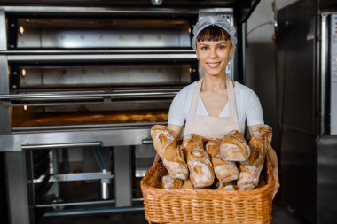 Kafkasyalı genç fırıncı fırında büyük bir sepet dolusu taze ekmek tutuyor. Elektrikli fırınların olduğu bir arka plan. Ekmek fırını fabrikası kavramı. Gülümseyen mutlu kadın kameraya bakıyor.