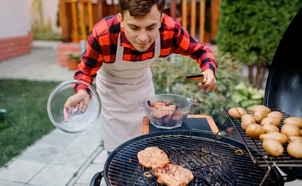穿着衬衫和围裙的年轻人正在自家花园的烤架上烤烤肉 — 图库照片