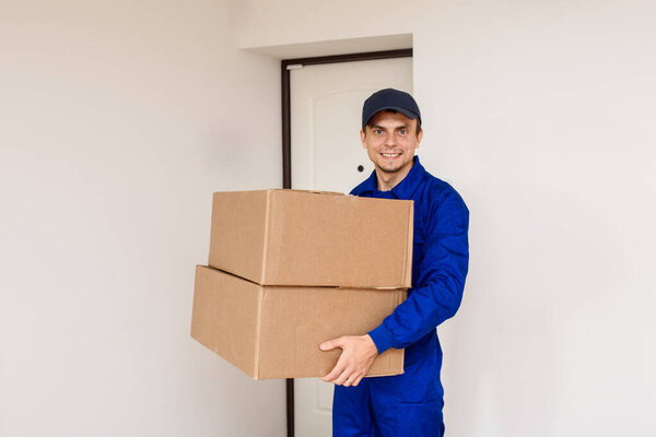 Young happy smiling man courier in blue overalls is delivering a parcel in two cardboard boxes. White background with door