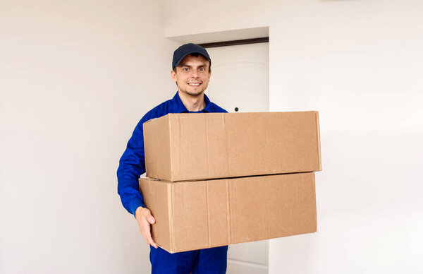 Young happy smiling man courier in blue overalls is delivering a parcel in two cardboard boxes. White background with door