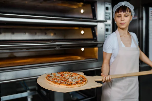 Młoda Biała Kobieta Piekarz Trzyma Skórkę Drewna Świeżą Pizzą Pobliżu — Zdjęcie stockowe