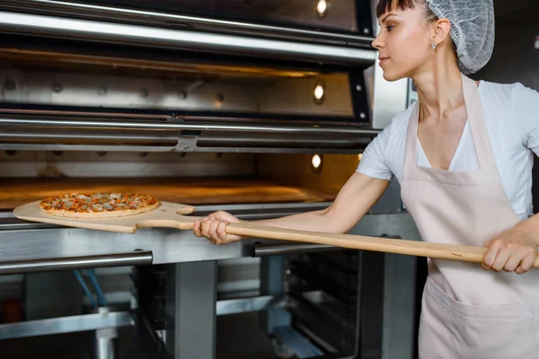 Young caucasian woman baker is holding a wood peel with fresh pizza and put it in an oven at a baking manufacture factory. Tasty pizza  factory concept.