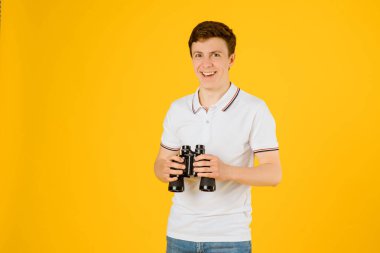 Beyaz tişörtlü, gülümseyen ve sarı arka plandaki dürbünle bakan mutlu bir adamın portresi. Spyglass turizm konsepti. Metnin için boşluk kopyala