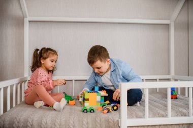 İki çocuk erkek ve kız kardeş Montessori yatağında oturmuş oyuncak tuğla küpleriyle oynuyorlar. Ev şeklindeki çocuklar için yatak.