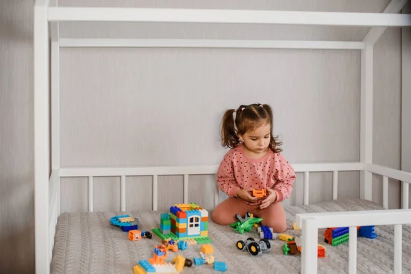 女の子はモンテッソーリのベッドに座っておもちゃのレンガキューブで遊んでいます 家の形をした子供用ベッド — ストック写真
