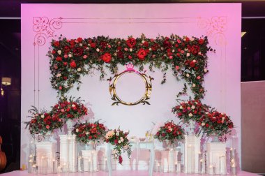 Düğün fotoğrafı standı kırmızı şakayık ve güllerle süslenmiş. Beyaz vazoda çiçekler. Şeffaf gözlüklerdeki mumlar