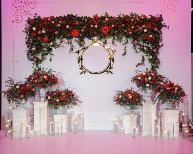 Düğün fotoğrafı standı kırmızı şakayık ve güllerle süslenmiş. Beyaz vazoda çiçekler. Şeffaf gözlüklerdeki mumlar