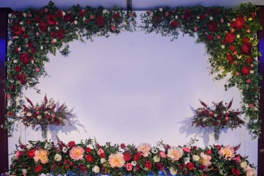 Düğün fotoğrafı standı kırmızı şakayık ve güllerle süslenmiş. Beyaz vazoda çiçekler. Şeffaf gözlüklerdeki mumlar