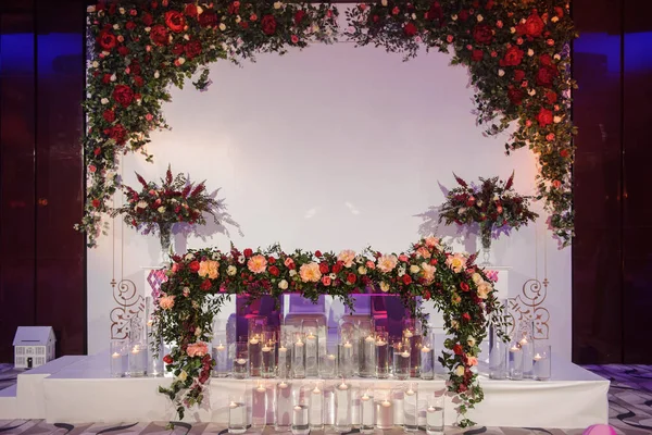 Düğün fotoğrafı standı kırmızı şakayık ve güllerle süslenmiş. Beyaz vazoda çiçekler. Şeffaf gözlüklerdeki mumlar