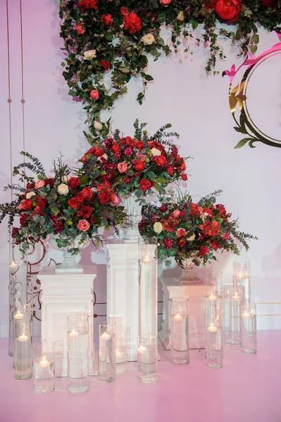 Düğün fotoğrafı standı kırmızı şakayık ve güllerle süslenmiş. Beyaz vazoda çiçekler. Şeffaf gözlüklerdeki mumlar