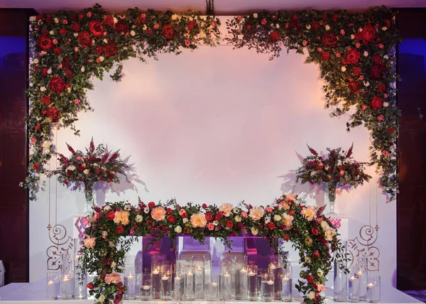 Düğün fotoğrafı standı kırmızı şakayık ve güllerle süslenmiş. Beyaz vazoda çiçekler. Şeffaf gözlüklerdeki mumlar