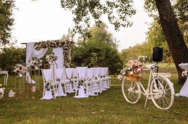 Park / Orman 'da bir düğün hazırlığı. Ahşap kemer ve sandalyeler beyaz ve pembe çiçeklerle süslenir güller ve şakayıklar, yerde bir sürü ağaç fener, mumlar, beyaz klasik bisiklet