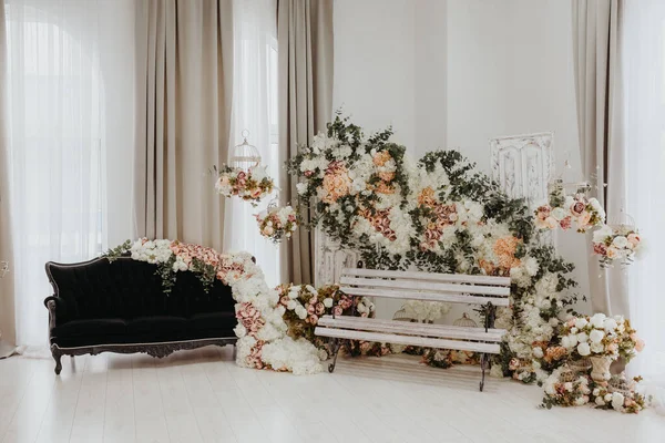 スタジオのヴィンテージウェディングパーティーフォトブースゾーンは 花の下に黒いソファと白い木のベンチで飾られています ストックフォト