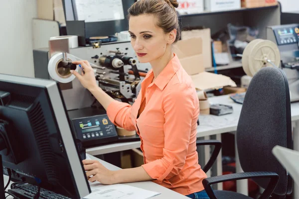 コンピュータ画面を見てワークショップでラベルプリンタに印刷されるラベルを設計する女性 — ストック写真