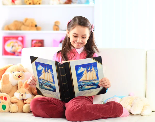 Encantadora niña leyendo libro con su juguete en un sofá blanco —  Fotos de Stock