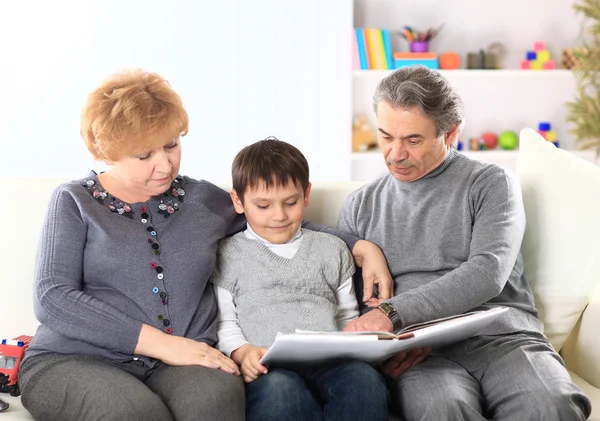 Cute boy sitting in the lap of their grandparents and looking happily together at a photo — Stock Photo, Image