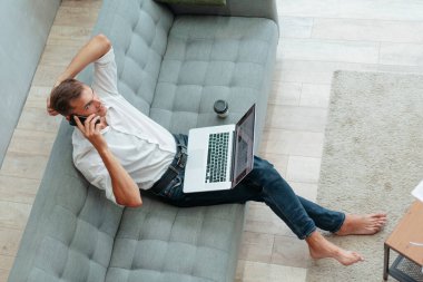 Genç adam akıllı bir telefonla konuşuyor, kanepede oturuyor..