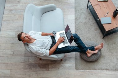 Kanepede otururken elinde bir fincan kahveyle dizüstü bilgisayar kullanan bir adam.