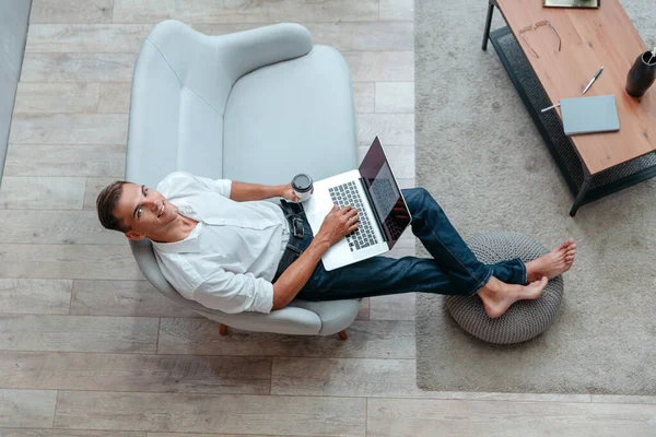 Muž s šálkem kávy pomocí notebooku, zatímco sedí na pohovce — Stock fotografie