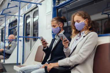 Metro vagonunda otururken akıllı telefonlarını kullanarak koruyucu maskeler takan yolcular..