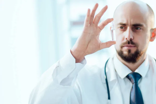 Científico sentado frente a un portátil abierto y mirando una ampolla con una vacuna. — Foto de Stock