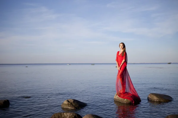 Uma jovem menina em um vestido vermelho em pé na costa — Fotografia de Stock