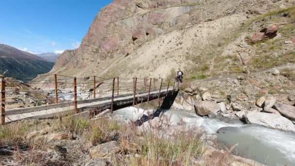 Mountaineer que cruza uma ponte pequena sobre uma corrente glacial alta da montanha — Vídeo de Stock