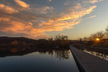 Tepelerde gün batımı gölün sularına yansıyor. Kıyılarında ve teknesinde bir kulübe var.