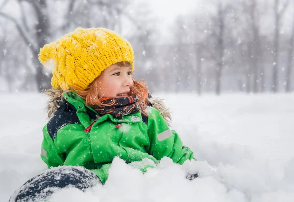 A happy baby girl is sitting in the snow and smiling. A little girl in a winter park during a snowfall. — Stock Photo, Image