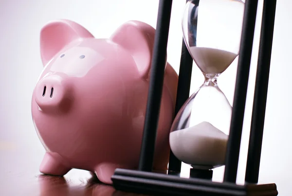 Pink piggy bank with a vintage hourglass behind — Stock Photo, Image