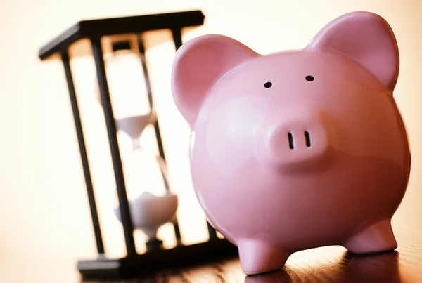 Pink piggy bank with a vintage hourglass behind — Stock Photo, Image