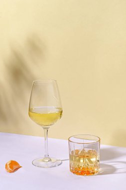 Sanatsal natürmort hayatı, iki farklı kadeh beyaz şarapla, açık havada beyaz bir masanın üzerinde, fotokopi için sarı duvara yaslanmış bir portakal diliminin yanında.