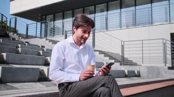 사업가가 커피를 마시고 벤치에 앉아 휴대 전화를 사용하는 모습 — 비디오