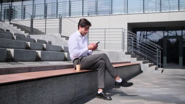 Empresario usando teléfono móvil y tomando café sentado en el banco — Vídeo de stock