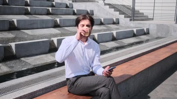 Az üzletember kávét iszik és mobiltelefont használ a padon ülve. — Stock videók